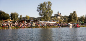 Crowd gathered along a riverbank with a castle in the distance.