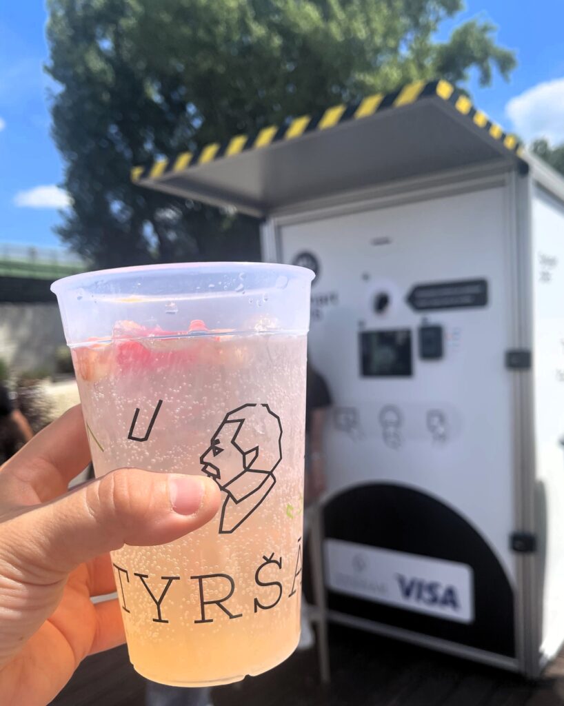 Hand holding a reusable Tyršák drink cup in front of a Smart DRS return station